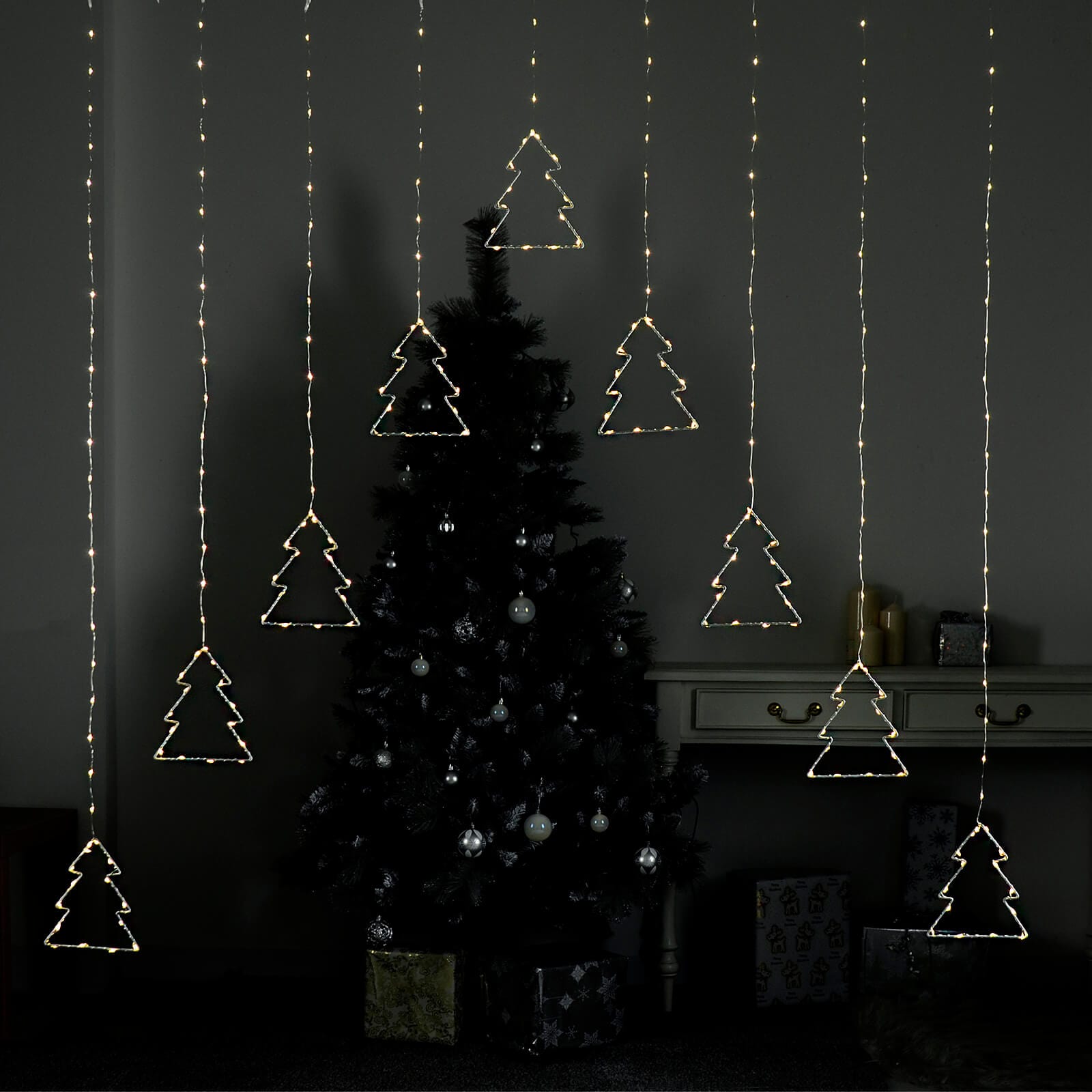 Christmas curtain light with 9 Christmas tree shapes lit by warm white LED lights, hanging in front of a Christmas tree and presents